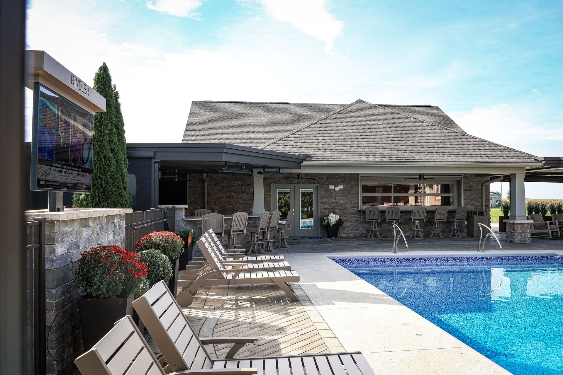 Backyard pool area with lounge chairs, modern stone and brick poolhouse with covered patio seating, manicured landscaping, and clear blue water on sunny day