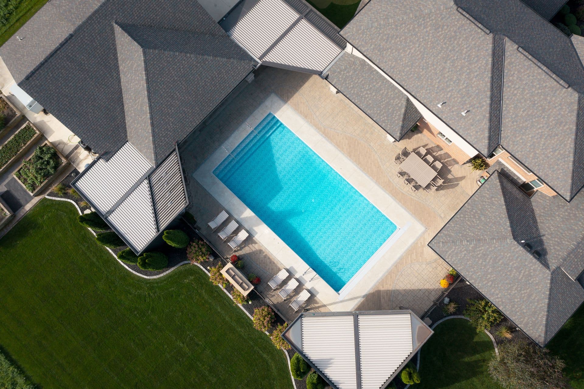 Aerial view of a residential swimming pool surrounded by lounge chairs and a white deck, nestled between multiple houses with gray shingled roofs