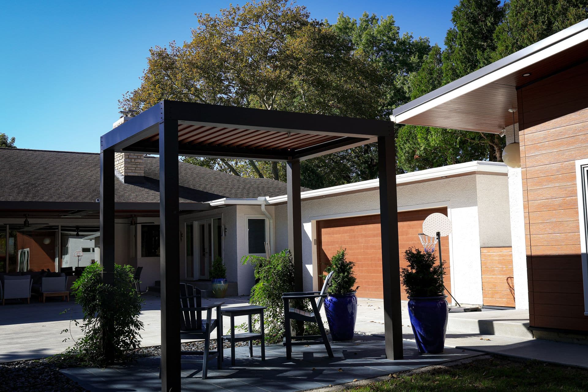 Modern backyard patio with black pergola structure over seating area, flanked by contemporary home with wood and white exterior, potted plants, and trees in background