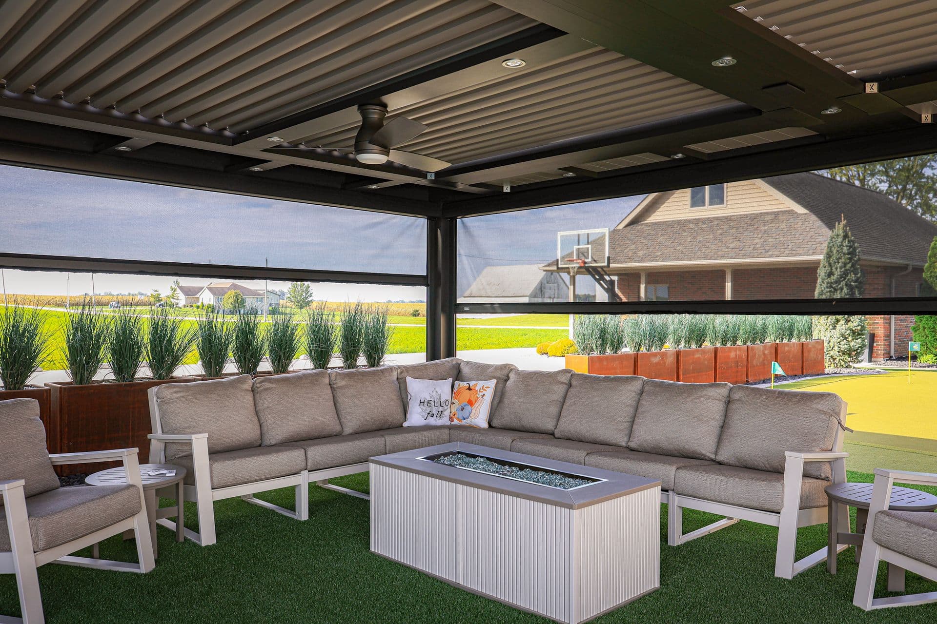 Modern covered patio with L-shaped sectional sofa, fire pit table, and lounge chairs overlooking residential backyard and open farmland