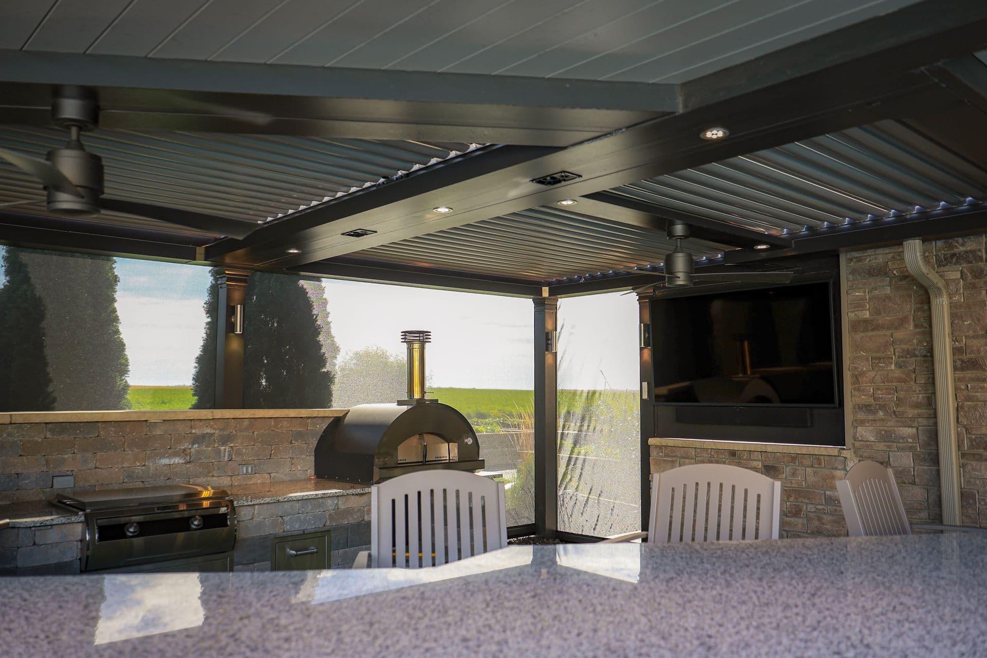 Outdoor kitchen and dining area with motorized louvered pergola, pizza oven, grill, countertop, chairs, and television mounted on brick wall overlooking green landscape