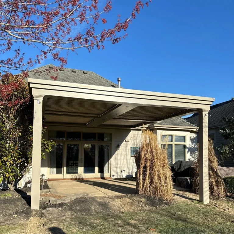 Backyard patio with a beige louvered pergola by Altimate Outdoors, installed over a concrete slab and attached to a modern home, surrounded by ornamental grasses and seasonal landscaping under a clear blue sky.